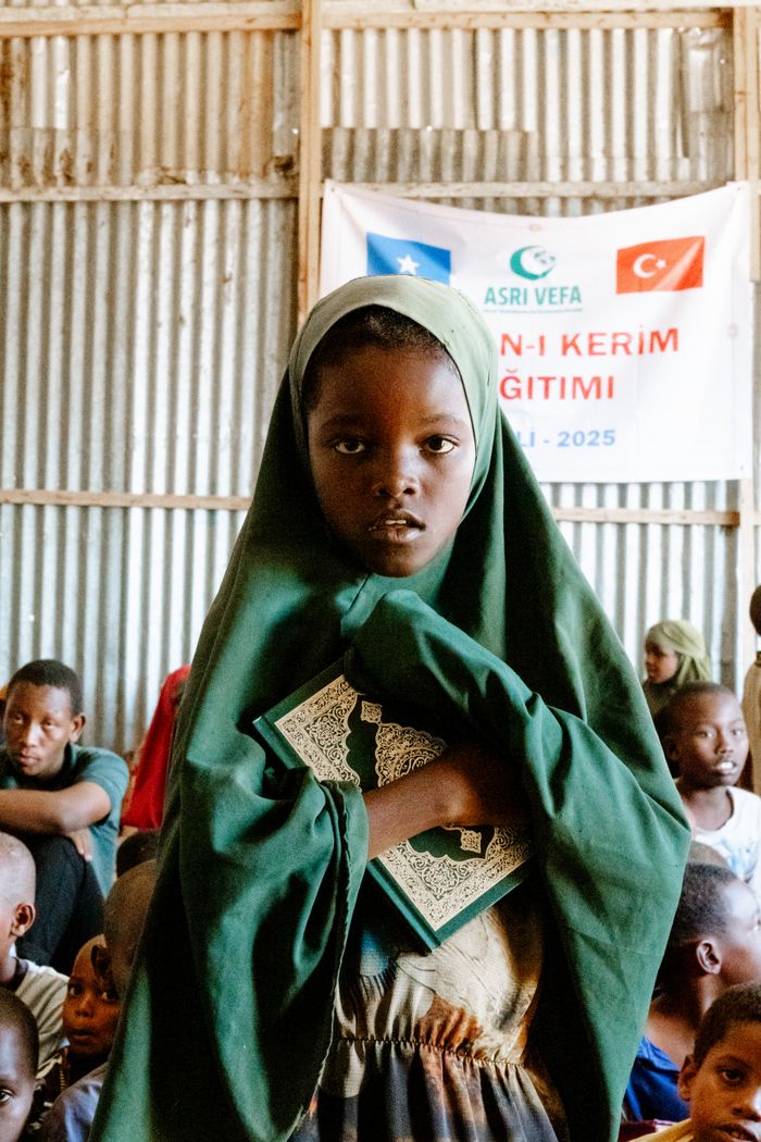 A young girl in a green hijab holds a religious book in an educational setting.