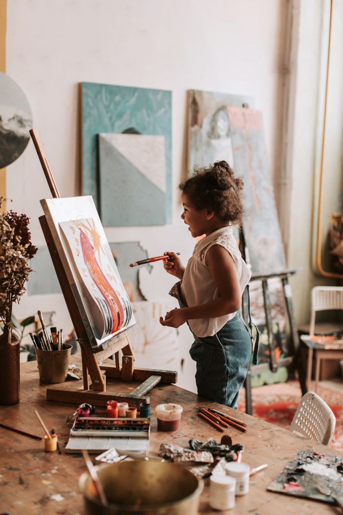 Home A young girl painting on a canvas indoors, surrounded by art supplies and canvases.